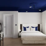 Nowoczesna sypialnia z białym łóżkiem kontynentalnym, drewnianą podłogą i granatowym sufitem, urządzona w minimalistycznym stylu z biurkiem, wieszakiem na ubrania oraz dużym oknem z jasnymi zasłonami, eleganckie wnętrze inspirowane granatową sypialnią